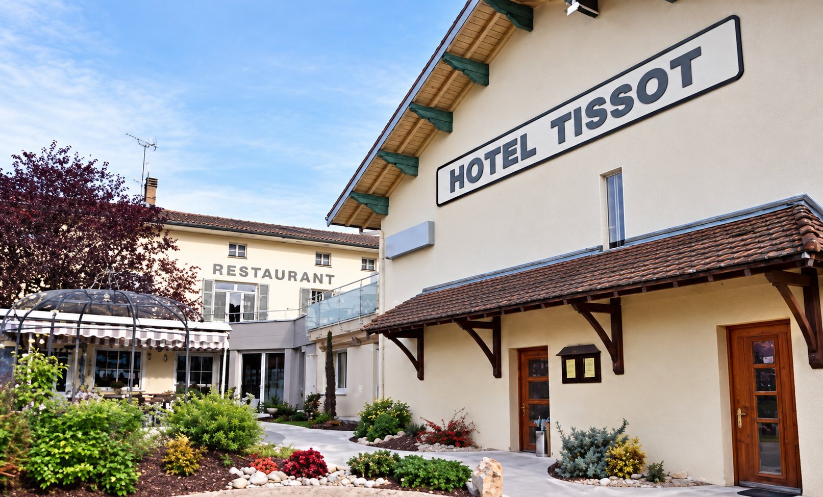 Façade de l’Hôtel Tissot avec son enseigne en biais, et le restaurant derrière les massifs de fleurs